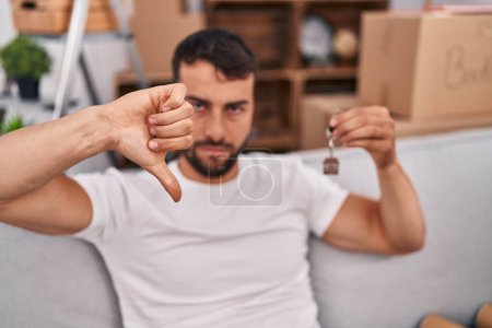 Foto de Hombre hispano guapo sosteniendo las llaves de un nuevo hogar con la cara enojada, signo negativo que muestra aversión con los pulgares hacia abajo, concepto de rechazo - Imagen libre de derechos