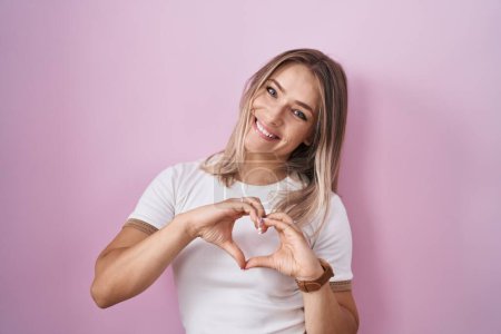 Téléchargez les photos : Blonde caucasian woman standing over pink background smiling in love doing heart symbol shape with hands. romantic concept. - en image libre de droit