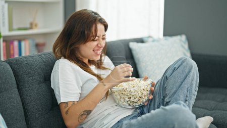 Téléchargez les photos : Jeune belle femme hispanique regardant un film manger du pop-corn assis sur un canapé à la maison - en image libre de droit