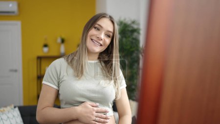 Téléchargez les photos : Jeune femme enceinte souriante confiante regardant ventre sur miroir à la maison - en image libre de droit