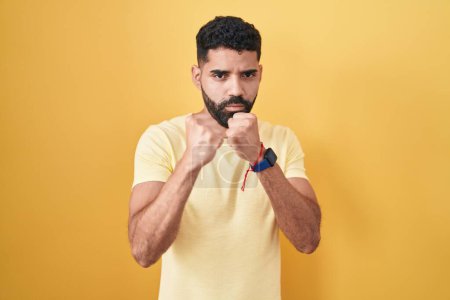 Téléchargez les photos : Homme hispanique avec barbe debout sur fond jaune prêt à se battre avec geste de défense poing, visage en colère et bouleversé, peur du problème - en image libre de droit