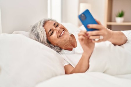 Téléchargez les photos : Femme d'âge moyen utilisant smartphone couché sur le lit dans la chambre - en image libre de droit