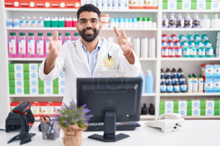 Téléchargez les photos : Homme hispanique avec barbe travaillant à la pharmacie pharmacie en regardant la caméra souriant à bras ouverts pour étreindre. expression joyeuse embrassant le bonheur. - en image libre de droit