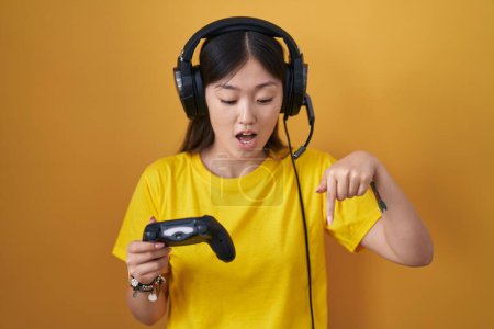 Foto de Chino joven mujer jugando videojuego celebración controlador apuntando hacia abajo con los dedos mostrando publicidad, cara sorprendida y boca abierta - Imagen libre de derechos