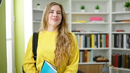 Téléchargez les photos : Jeune étudiante blonde souriante confiante tenant des livres à la bibliothèque de l'université - en image libre de droit