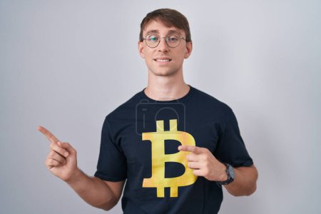 Foto de Hombre rubio caucásico con camiseta bitcoin sonriendo y mirando a la cámara apuntando con dos manos y dedos hacia un lado. - Imagen libre de derechos