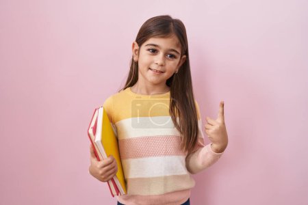 Photo for Little hispanic girl holding books smiling with an idea or question pointing finger with happy face, number one - Royalty Free Image
