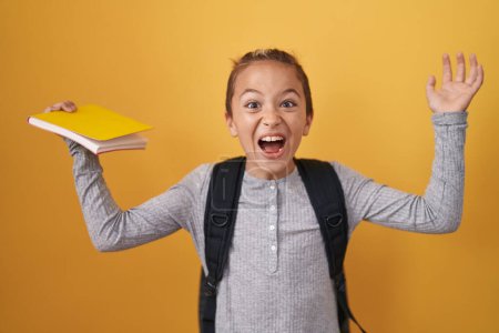 Photo for Little caucasian boy wearing student backpack and holding book celebrating victory with happy smile and winner expression with raised hands - Royalty Free Image