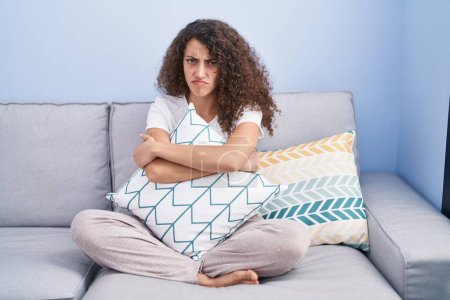 Foto de Mujer hispana con el pelo rizado sentada en el sofá en casa escéptica y nerviosa, frunciendo el ceño molesto debido a un problema. persona negativa. - Imagen libre de derechos