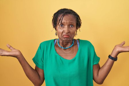 Foto de Mujer africana con rastas de pie sobre fondo amarillo expresión despistada y confusa con los brazos y las manos levantadas. concepto de duda. - Imagen libre de derechos