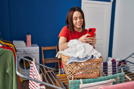 Photo for Young beautiful hispanic woman using smartphone hanging clothes on clothesline at laundry room - Royalty Free Image