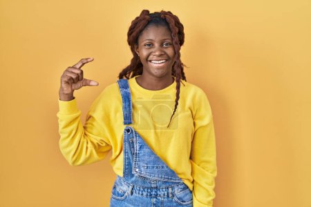 Téléchargez les photos : Femme africaine debout sur fond jaune geste souriant et confiant avec la main faisant signe de petite taille avec les doigts regardant et la caméra. concept de mesure. - en image libre de droit