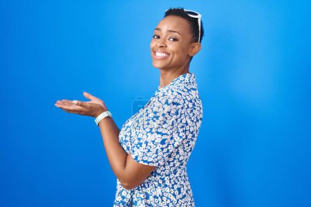Foto de Mujer afroamericana de pie sobre fondo azul señalando a un lado con las manos abiertas palmeras mostrando espacio de copia, presentando anuncio sonriendo emocionado feliz - Imagen libre de derechos