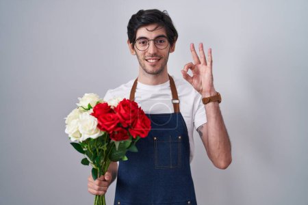 Foto de Joven hombre hispano sosteniendo ramo de rosas blancas y rojas sonriendo positiva haciendo signo de ok con la mano y los dedos. expresión exitosa. - Imagen libre de derechos