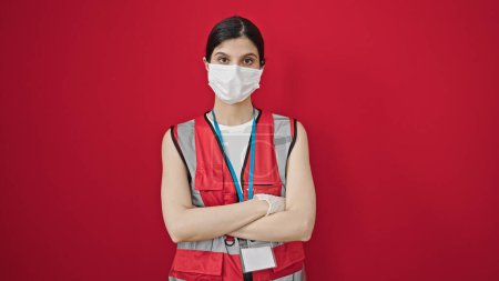 Foto de Joven hermosa doctora hispana con máscara médica de pie con los brazos cruzados gesto sobre fondo rojo aislado - Imagen libre de derechos