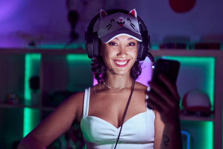 Téléchargez les photos : Jeune belle hispanique femme streamer souriant confiant en utilisant smartphone à la salle de jeu - en image libre de droit