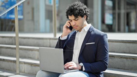 Photo for Young latin man business worker using laptop talking on smartphone at street - Royalty Free Image