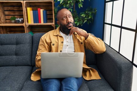 Foto de Hombre afroamericano usando portátil en casa sentado en el sofá cara seria pensando en la pregunta con la mano en la barbilla, reflexivo acerca de la idea confusa - Imagen libre de derechos