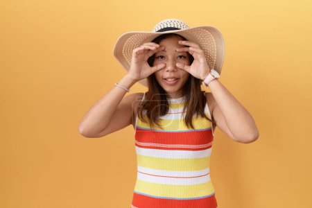 Téléchargez les photos : Femme chinoise d'âge moyen portant un chapeau d'été sur fond jaune essayant d'ouvrir les yeux avec les doigts, endormie et fatiguée pour la fatigue matinale - en image libre de droit
