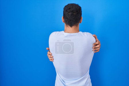 Foto de Joven hombre hispano de pie sobre fondo azul abrazándose feliz y positivo desde atrás. amor propio y cuidado personal - Imagen libre de derechos