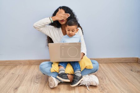 Téléchargez les photos : Jeune mère hispanique et enfant utilisant ordinateur portable assis sur le sol couvrant les yeux avec la main, l'air grave et triste. concept de non-visibilité, de dissimulation et de rejet - en image libre de droit