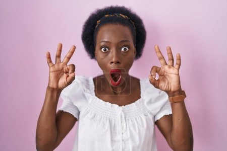 Photo for African woman with curly hair standing over pink background looking surprised and shocked doing ok approval symbol with fingers. crazy expression - Royalty Free Image