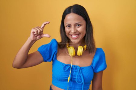 Téléchargez les photos : Jeune femme hispanique debout sur fond jaune geste souriant et confiant avec la main faisant signe de petite taille avec les doigts regardant et la caméra. concept de mesure. - en image libre de droit