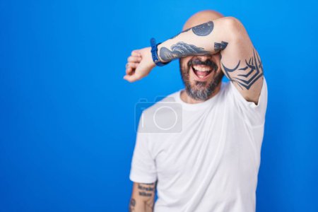Foto de Hombre hispano con tatuajes de pie sobre fondo azul cubriendo los ojos con el brazo sonriendo alegre y divertido. concepto ciego. - Imagen libre de derechos
