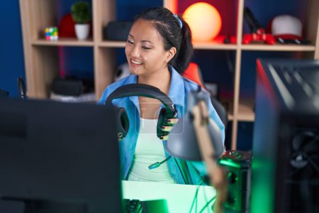Foto de Joven mujer china streamer sonriendo con confianza celebración de auriculares en la sala de juegos - Imagen libre de derechos