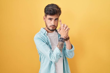 Téléchargez les photos : Jeune homme hispanique avec des tatouages debout sur fond jaune tenant arme symbolique avec geste de la main, jouant tuer des armes de tir, visage en colère - en image libre de droit