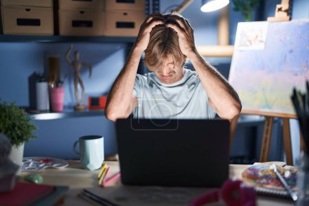 Téléchargez les photos : Homme d'âge moyen assis au studio d'art avec ordinateur portable la nuit souffrant de maux de tête désespérés et stressés parce que la douleur et la migraine. mains sur la tête. - en image libre de droit