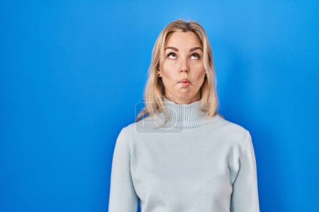Foto de Young caucasian woman standing over blue background making fish face with lips, crazy and comical gesture. funny expression. - Imagen libre de derechos