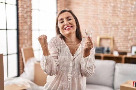 Photo for Middle age hispanic woman holding lightbulb for inspiration and idea screaming proud, celebrating victory and success very excited with raised arms - Royalty Free Image