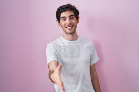 Foto de Joven hombre hispano de pie sobre fondo rosa sonriente amistoso ofreciendo apretón de manos como saludo y bienvenida. negocios exitosos. - Imagen libre de derechos