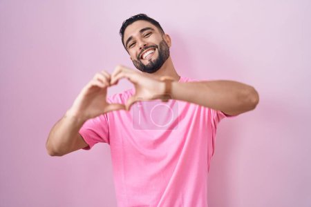 Téléchargez les photos : Jeune homme hispanique debout sur fond rose souriant dans l'amour faisant forme de symbole de coeur avec les mains. concept romantique. - en image libre de droit