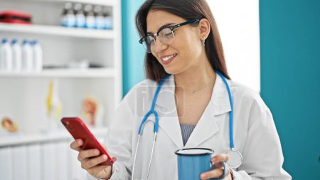 Téléchargez les photos : Jeune belle hispanique femme médecin en utilisant smartphone boire du café à la clinique - en image libre de droit