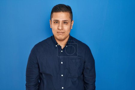 Photo for Hispanic young man standing over blue background relaxed with serious expression on face. simple and natural looking at the camera. - Royalty Free Image