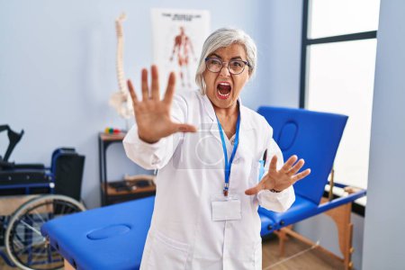 Photo for Middle age woman with grey hair working at pain recovery clinic afraid and terrified with fear expression stop gesture with hands, shouting in shock. panic concept. - Royalty Free Image