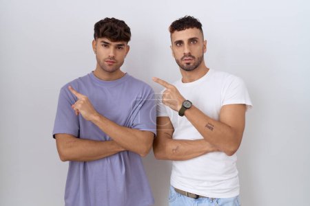 Téléchargez les photos : Homosexuel gay couple debout sur fond blanc pointant avec doigt de la main sur le côté montrant la publicité, sérieux et calme visage - en image libre de droit