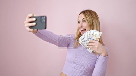 Foto de Joven mujer hispana hermosa tomando selfie sosteniendo dólares sobre fondo rosa aislado - Imagen libre de derechos