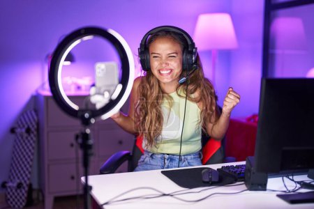 Téléchargez les photos : Jeune femme hispanique jouant à des jeux vidéo enregistrement avec smartphone criant fier, célébrant la victoire et le succès très excité avec bras levé - en image libre de droit