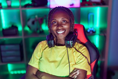 Foto de Mujer afroamericana streamer sonriendo confiado sentado con los brazos cruzados gesto en sala de juegos - Imagen libre de derechos