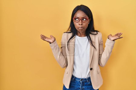 Photo for African young woman wearing glasses clueless and confused expression with arms and hands raised. doubt concept. - Royalty Free Image