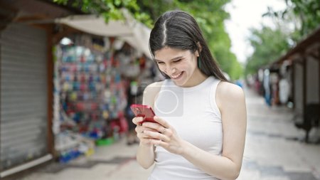 Foto de Mujer hispana hermosa joven usando teléfono inteligente sonriendo en el mercado callejero - Imagen libre de derechos