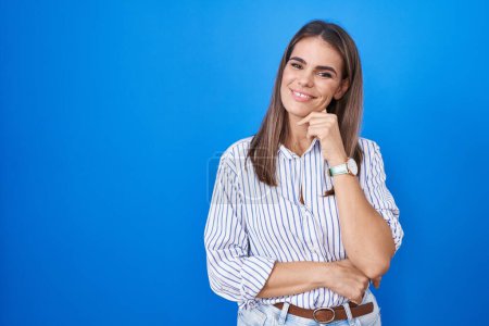 Foto de Hispanic young woman standing over blue background looking confident at the camera smiling with crossed arms and hand raised on chin. thinking positive. - Imagen libre de derechos