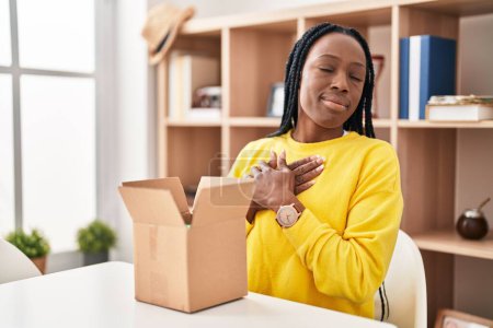 Photo for Beautiful black woman opening cardboard box smiling with hands on chest, eyes closed with grateful gesture on face. health concept. - Royalty Free Image