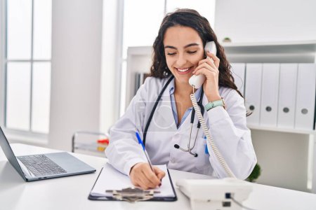 Téléchargez les photos : Jeune femme hispanique portant l'uniforme de médecin parlant au téléphone à la clinique - en image libre de droit