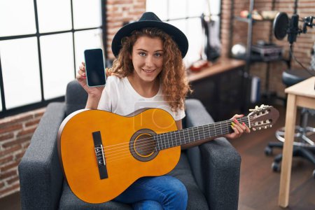 Téléchargez les photos : Jeune femme caucasienne jouant de la guitare classique au studio de musique tenant smartphone souriant avec un sourire heureux et cool sur le visage. montrant les dents. - en image libre de droit