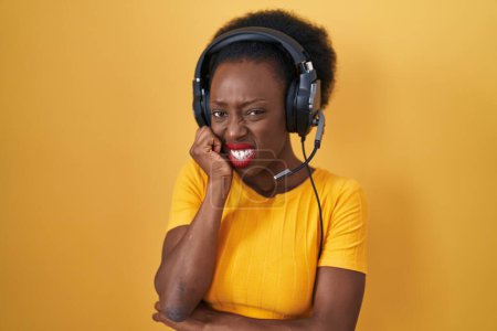 Photo for African woman with curly hair standing over yellow background wearing headphones looking stressed and nervous with hands on mouth biting nails. anxiety problem. - Royalty Free Image