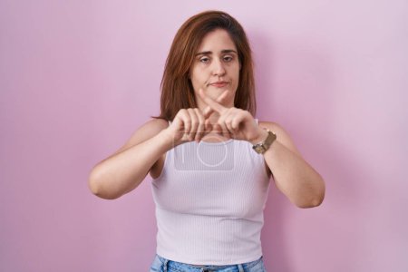 Photo for Brunette woman standing over pink background rejection expression crossing fingers doing negative sign - Royalty Free Image